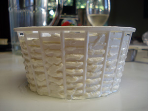 a ricotta-form with ricotta squeezing through the basket mesh and a white wine glass behind