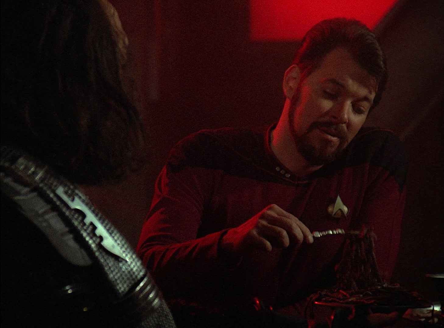 Commander Riker in his Starfleet uniform considers eating a bite of live serpent worms while talking to his Klingon crewmate during an officer exchange